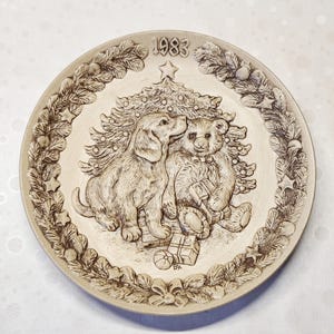 Könnte beinhalten: Ein runder, cremefarbener Zierteller mit einem erhabenen, einfarbigen Reliefdesign, das einen Welpen und einen Teddybären vor einem Weihnachtsbaum zeigt. Der Rand ist mit Tannenzweigen verziert und die Jahreszahl 1983 ist oben eingeprägt.