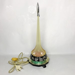 May include: A vintage rocket-shaped lamp with a silver tip and a clear, yellow-tinted body. The base is black with green and pink accents, and the lamp has a power cord with a yellow plug. The lamp is a retro space-themed decorative item.