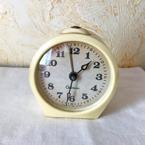 Pode incluir: Um despertador vintage branco com números e ponteiros pretos. O mostrador do relógio diz "Слава" e "СССР".
