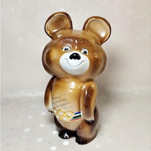 Puede incluir: Una figura de oso de cerámica marrón brillante con orejas grandes y redondeadas, hocico blanco y nariz negra. El oso lleva un cinturón con los cinco anillos olímpicos entrelazados en oro, azul, negro, verde y rojo, con texto cirílico tenue.