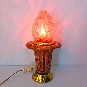 May include: A ceramic table lamp with a brown base and a clear glass shade shaped like a flame. The base features a design of figures dancing around a pole. The lamp is lit and the light shines through the glass shade.