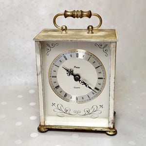 May include: A rectangular mantel clock with a distressed white finish and a gold-toned handle on top. The clock face features black Roman numerals, the brand name Paico, the word Quartz, and decorative black scrollwork above and below the dial.