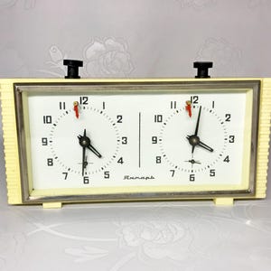 May include: A vintage white chess clock with two separate timers, each with black hands and red second markers. The clock has a cream-coloured face with black numbers and the word "Янтарь" printed on the bottom.