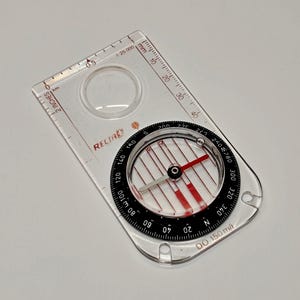May include: A clear rectangular base plate compass with a black rotating bezel featuring degree markings from 0 to 360. The device includes a red and silver needle, a magnifying lens, and printed scales in inches and millimeters with the text RECTA.