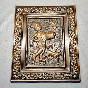 May include: A rectangular brass wall plaque featuring a relief design of a person playing an accordion with a small dog nearby. The central scene is surrounded by an ornate, embossed floral border with a polished finish and dark recessed details.