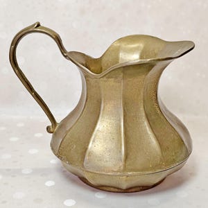 May include: A metallic gold-toned pitcher featuring a scalloped, fluted body design with a wide pouring spout and a curved handle. The surface has a matte finish with visible texture and subtle variations in the metal tone across the exterior.