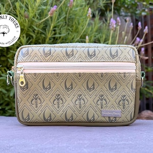 May include: Olive green crossbody bag with a repeating diamond pattern and a zipper closure. The bag features a gold zipper pull and a small tag that reads "The Starry Dreamer". The bag has the text "Cottonly Yours" on a circular tag.
