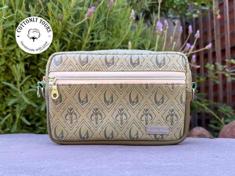 May include: Olive green crossbody bag with a repeating diamond pattern and a zipper closure. The bag features a gold zipper pull and a small tag that reads "The Starry Dreamer". The bag has the text "Cottonly Yours" on a circular tag.