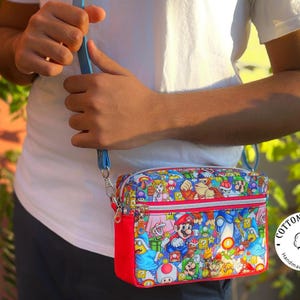 May include: A colorful crossbody bag featuring a vibrant print of various characters from the Super Mario video game series. The bag has a red base, a blue strap, and a zipper closure. The bag is held by a person wearing a white t-shirt.
