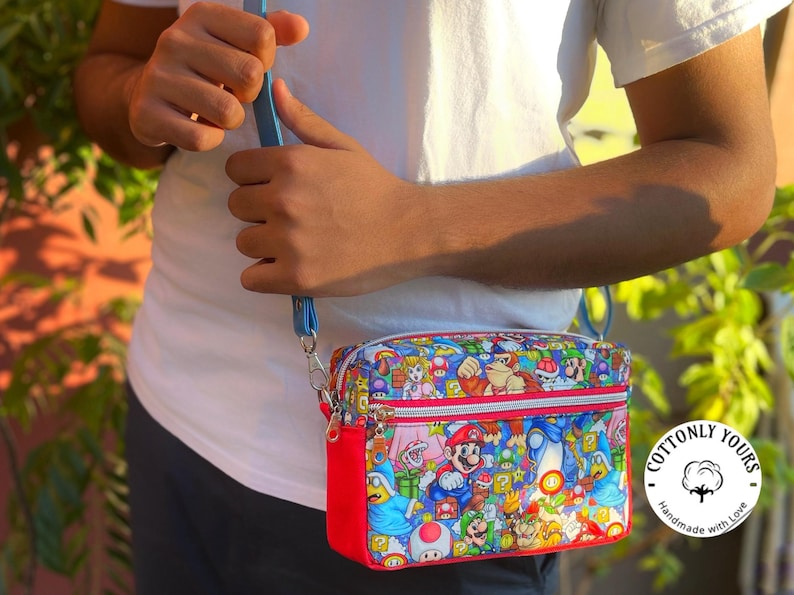 May include: A colorful crossbody bag featuring a vibrant print of various characters from the Super Mario video game series. The bag has a red base, a blue strap, and a zipper closure. The bag is held by a person wearing a white t-shirt.
