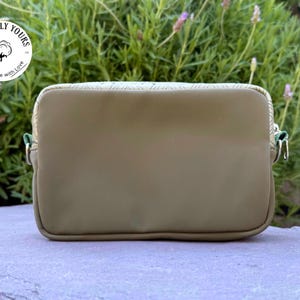 May include: Olive green rectangular crossbody bag with a zipper closure and silver-toned hardware. The bag has a textured top panel and a green zipper pull. The bag is sitting on a stone surface with a green plant background. The bag has the text "Cottonly Yours" on it.
