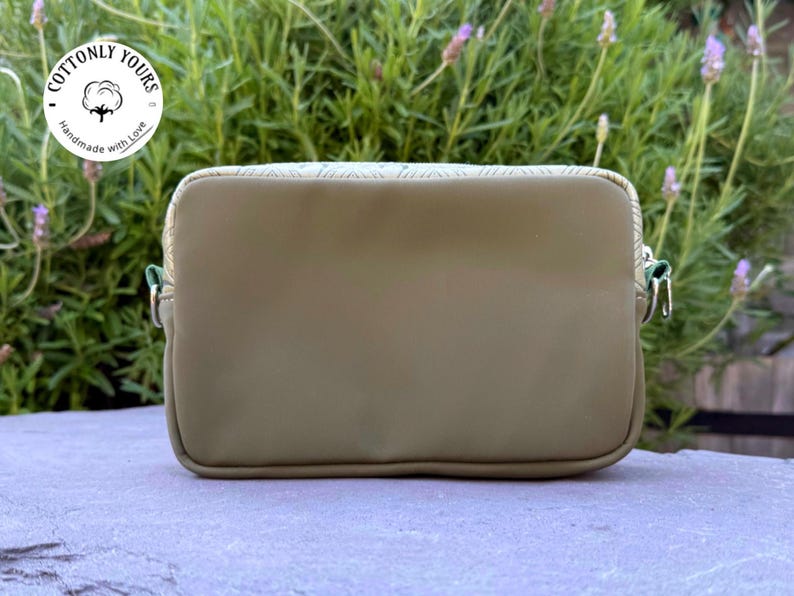 May include: Olive green rectangular crossbody bag with a zipper closure and silver-toned hardware. The bag has a textured top panel and a green zipper pull. The bag is sitting on a stone surface with a green plant background. The bag has the text "Cottonly Yours" on it.