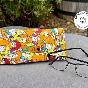 May include: A rectangular fabric glasses case featuring a repeating cartoon print of characters from Scooby-Doo, including Scooby-Doo, Shaggy, Velma, Daphne, and Fred, in orange, yellow, purple, and green, paired with black metal-framed eyeglasses.