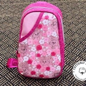 Puede incluir: Mochila bandolera rosa con estampado floral y de gatos. La bolsa tiene una sola correa ajustable y un compartimento principal con cremallera. El diseño incluye flores rosas, blancas y rojas, e ilustraciones de gatos de dibujos animados.