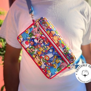 May include: A rectangular crossbody bag featuring a colorful print of Mario, Luigi, and other characters from the Super Mario Bros. video game series. The bag has a red trim, a silver zipper, and a blue strap. The bag is worn over the shoulder.
