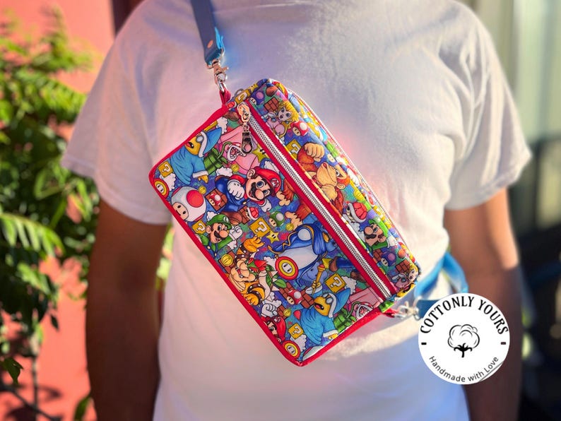 May include: A rectangular crossbody bag featuring a colorful print of Mario, Luigi, and other characters from the Super Mario Bros. video game series. The bag has a red trim, a silver zipper, and a blue strap. The bag is worn over the shoulder.