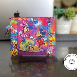 May include: A pink zippered pouch with a colorful print of Mickey Mouse, Minnie Mouse, Donald Duck, Goofy, and Pluto. The pouch has a purple flap and a silver zipper pull. The text "Cottonly Yours" is printed on a white circle with a cotton boll image. The text "Handmade with Love" is printed below the circle.