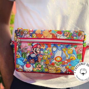 May include: A colorful fanny pack featuring a vibrant print of characters from the Super Mario video game series. The bag has a red zipper and trim, a blue strap, and the text "Cottonly Yours".