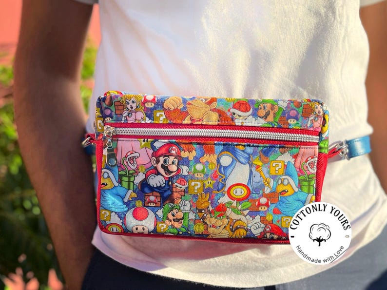 May include: A colorful fanny pack featuring a vibrant print of characters from the Super Mario video game series. The bag has a red zipper and trim, a blue strap, and the text "Cottonly Yours".