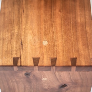 May include: Close-up of a wooden box with a light brown finish. The box has a decorative edge with a series of small, triangular pieces of wood. The top of the box has two small, round wooden plugs.
