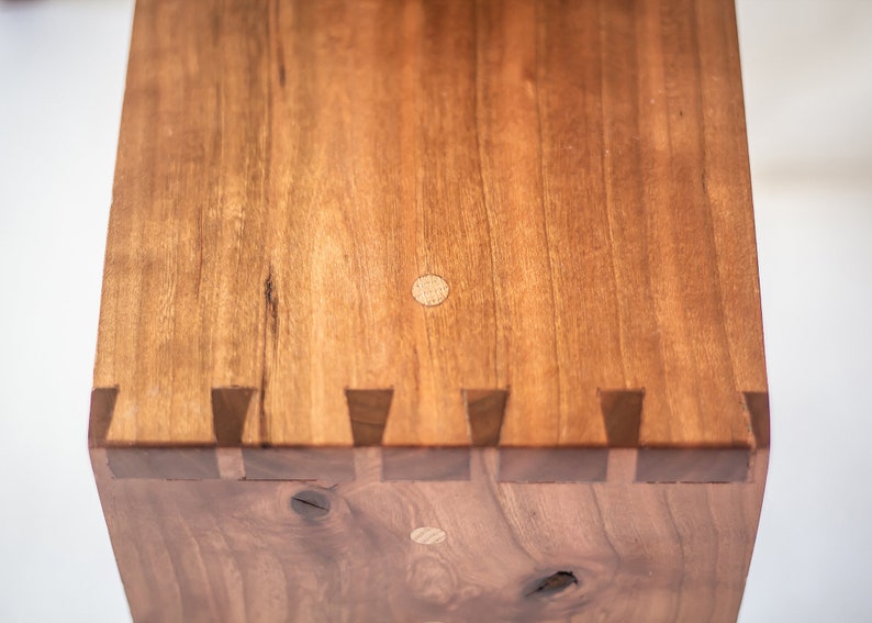 May include: Close-up of a wooden box with a light brown finish. The box has a decorative edge with a series of small, triangular pieces of wood. The top of the box has two small, round wooden plugs.