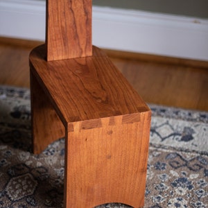 May include: A wooden stool with a simple design, featuring a rectangular seat and a tall back. The stool is made of cherry wood and has a natural finish.