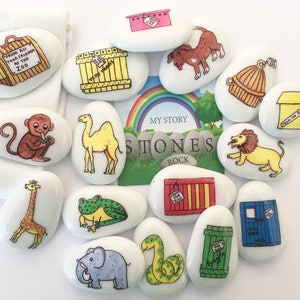 May include: A selection of painted white stones, each depicting a different zoo animal and crates. The illustrations include a monkey, camel, giraffe, frog, elephant, snake, and lion. The stones are arranged around a book with a rainbow and the words "My Story Stones Rock."