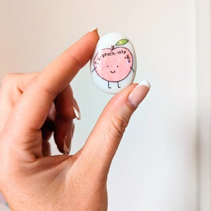 May include: A white painted rock with a peach design and the text "I-a-peach-iate you".