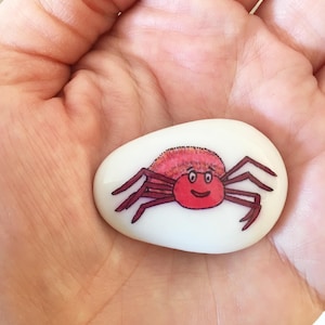 May include: A white painted rock with a red and pink crab painted on it. The crab has a smiling face and is facing the viewer.