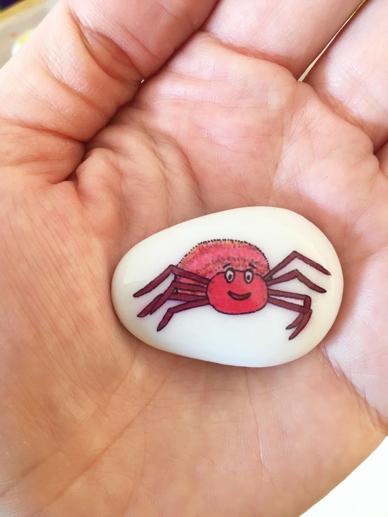 May include: A white painted rock with a red and pink crab painted on it. The crab has a smiling face and is facing the viewer.