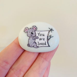 May include: White painted rock with a cartoon koala holding a sign that says "You are loved".