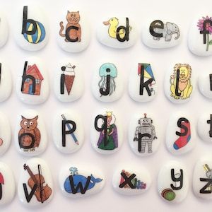 May include: Set of 26 white painted stones with black letters and colourful illustrations. Each stone features a different letter of the alphabet, from A to Z, with a corresponding image. The images include a cat, a duck, an elephant, a flower, grapes, a house, ice cream, a jellyfish, a kite, a lion, a mouse, a nose, an owl, a pencil, a queen, a robot, a sock, a train, an umbrella, a violin, a whale, a xylophone, a yo-yo, and a zipper.