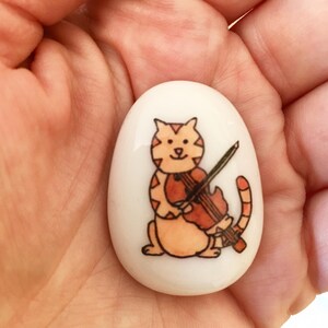 May include: A white ceramic stone with a hand-painted illustration of an orange tabby cat playing a violin. The cat has a black nose and black eyes.