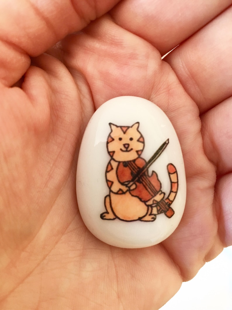 May include: A white ceramic stone with a hand-painted illustration of an orange tabby cat playing a violin. The cat has a black nose and black eyes.