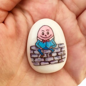 May include: A white ceramic egg-shaped stone with a hand-painted illustration of a cartoon character sitting on a brick wall. The character is wearing a blue shirt, red bowtie, and has a pink face.