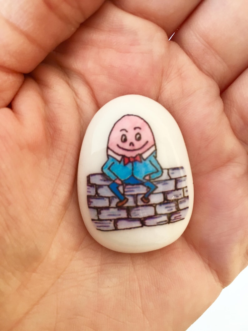 May include: A white ceramic egg-shaped stone with a hand-painted illustration of a cartoon character sitting on a brick wall. The character is wearing a blue shirt, red bowtie, and has a pink face.