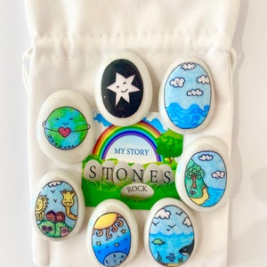 May include: Set of seven white oval stones with colorful illustrations. The stones feature a star, clouds, a globe, a giraffe, a sun, a turtle, and a tree. The stones are in a white drawstring bag with the words "My Story Stones Rock" printed on the bag.