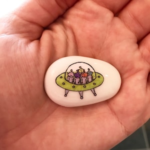 May include: A white painted rock with a green UFO and five colorful aliens inside. The aliens are purple, yellow, orange, green, and blue.