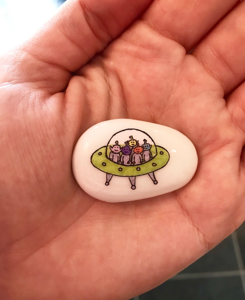 May include: A white painted rock with a green UFO and five colorful aliens inside. The aliens are purple, yellow, orange, green, and blue.