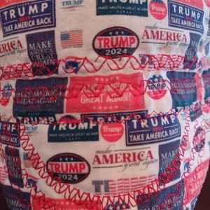 May include: A white fabric with a red, white, and blue pattern featuring the words "Trump" and "Make America Great Again". The fabric is sewn with red stitching.