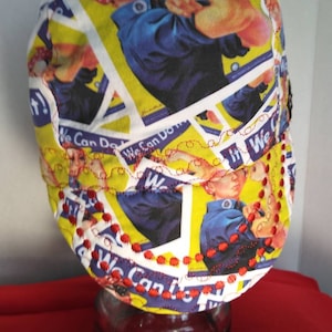 May include: A yellow, white and blue fabric hat with a red, white and blue image of a woman with her arm raised. The hat has red stitching around the edge and the words "We Can Do It!" are visible on the fabric.