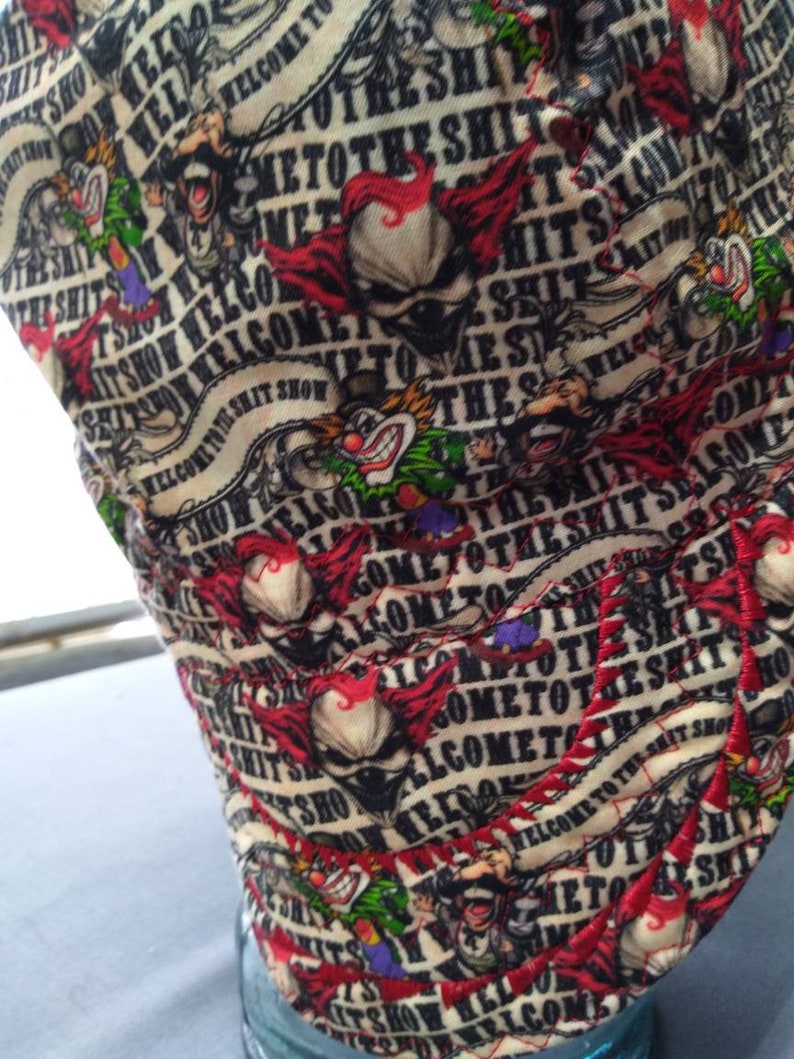 May include: A white fabric hat with a red, black, and green pattern. The hat has the text "Welcome to the Shit Show" repeated in a circular pattern.