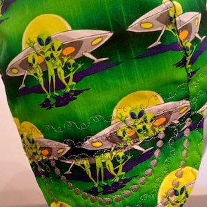 May include: A green welding cap with a repeating pattern of aliens in flying saucers. The cap has a decorative, quilted edge.
