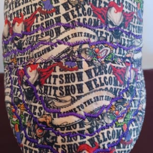 May include: A beige baseball cap with a colorful, cartoon-like design. The cap features the text "Welcome to the Shit Show" repeated in a circular pattern. The text is embroidered in purple thread.