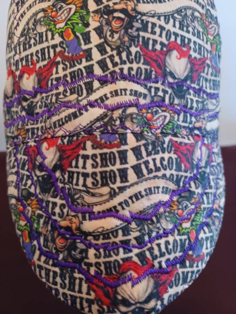May include: A beige baseball cap with a colorful, cartoon-like design. The cap features the text "Welcome to the Shit Show" repeated in a circular pattern. The text is embroidered in purple thread.