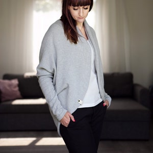 May include: A woman wearing a gray cardigan with a white shirt underneath and black pants. She is standing in a room with a couch in the background.