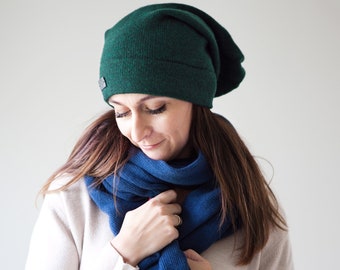 May include: A woman wearing a dark green beanie and a blue scarf. The scarf is wrapped around her neck and she is holding it with her right hand.
