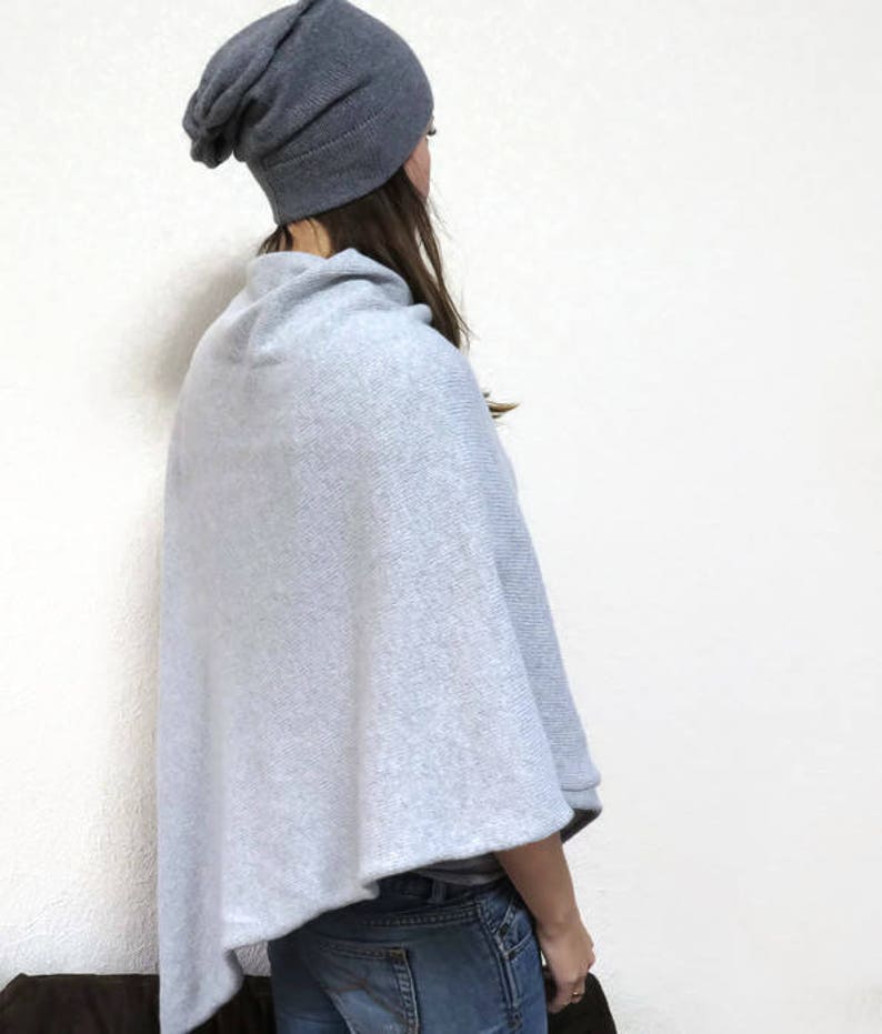 May include: A light gray knitted poncho with a slouchy gray beanie. The poncho has a cowl neck and is worn over a pair of blue jeans.