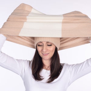 Op de afbeelding: Een vrouw draagt een wit shirt met lange mouwen en een beige en witte gestreepte gebreide sjaal over haar hoofd.