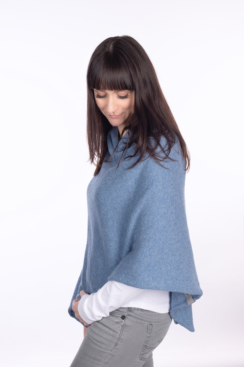 Pode incluir: Uma mulher vestindo um poncho de malha azul com uma camisa branca de manga comprida por baixo e cal&ccedil;as de ganga cinzas.